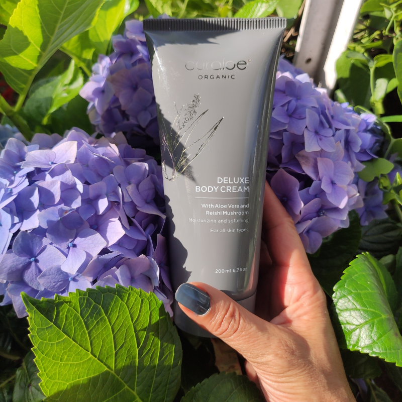Hand holding a tube of Organic Deluxe Body Cream against purple flowers