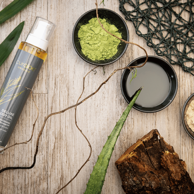 Curaloe Body oil bottle, green powder, and natural elements on a wooden surface