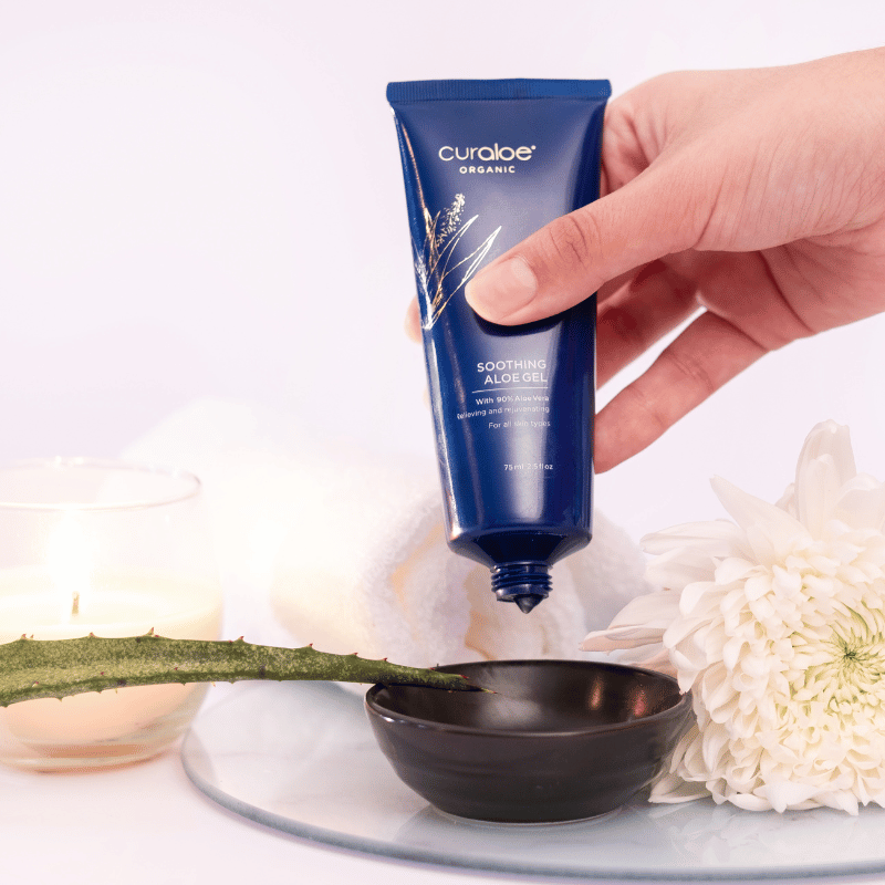 Hand holding a blue Curaloe aloe gel tube with flowers in the background