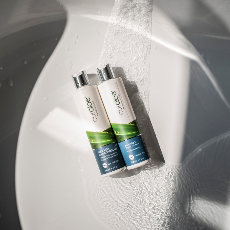 Two bottles of Curaloe products on a white surface with water droplets.