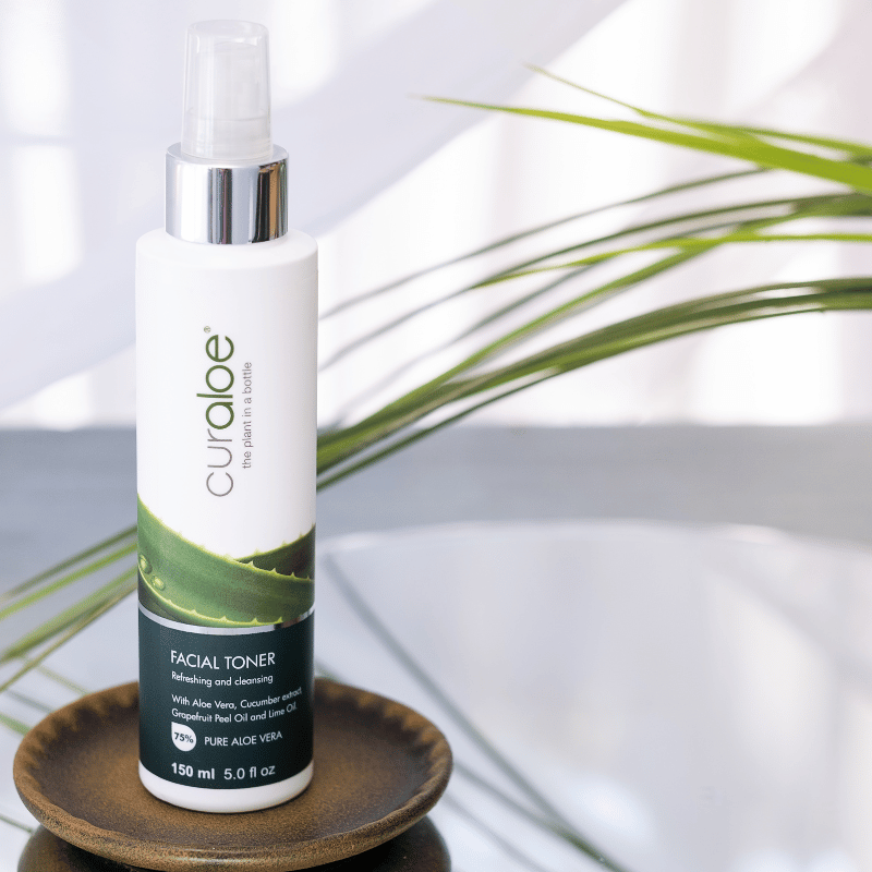 Curaloe facial toner bottle on a wooden coaster with a plant in the background