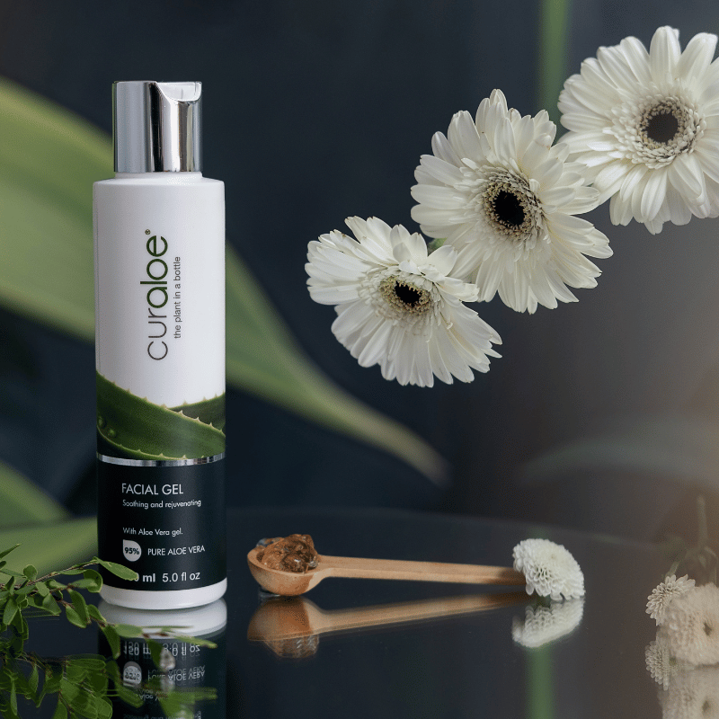 Curaloe facial gel bottle with white flowers and a wooden spoon on a reflective surface