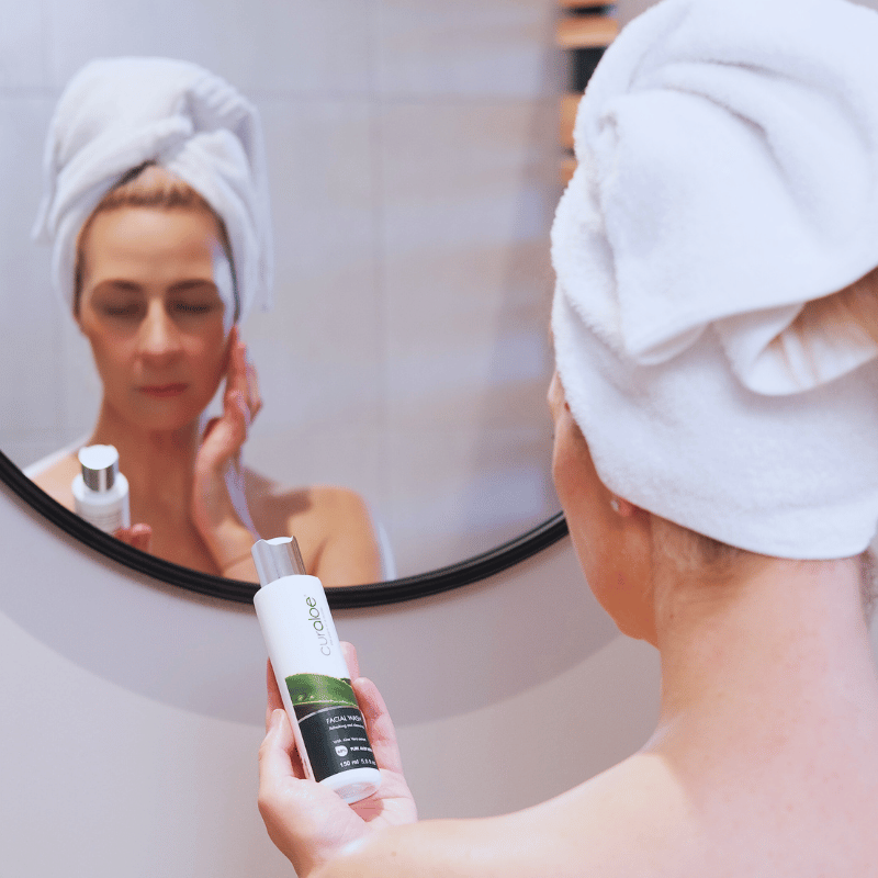 Woman applying Curaloe Facial Wash in front of a mirror with a towel on her head