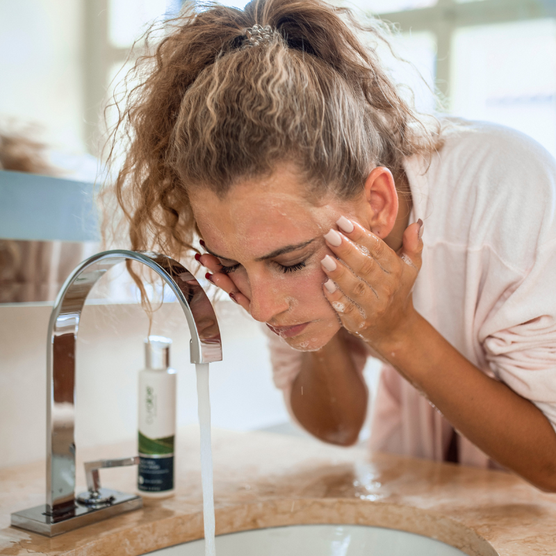 Woman washing her face at a bathroom sink with a bottle of Curaloe Facial Wash nearby.