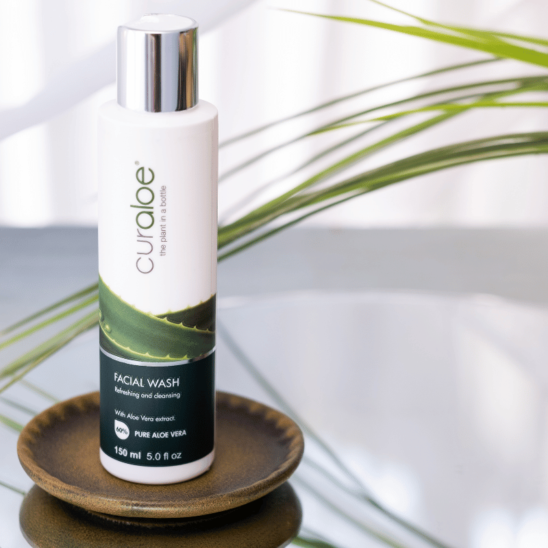 Curaloe facial wash bottle on a wooden coaster with green leaves in the background