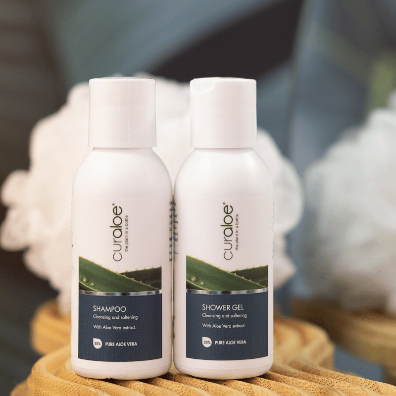 Two bottles of Curaloe shampoo and shower gel on a wooden surface with a blurred background.