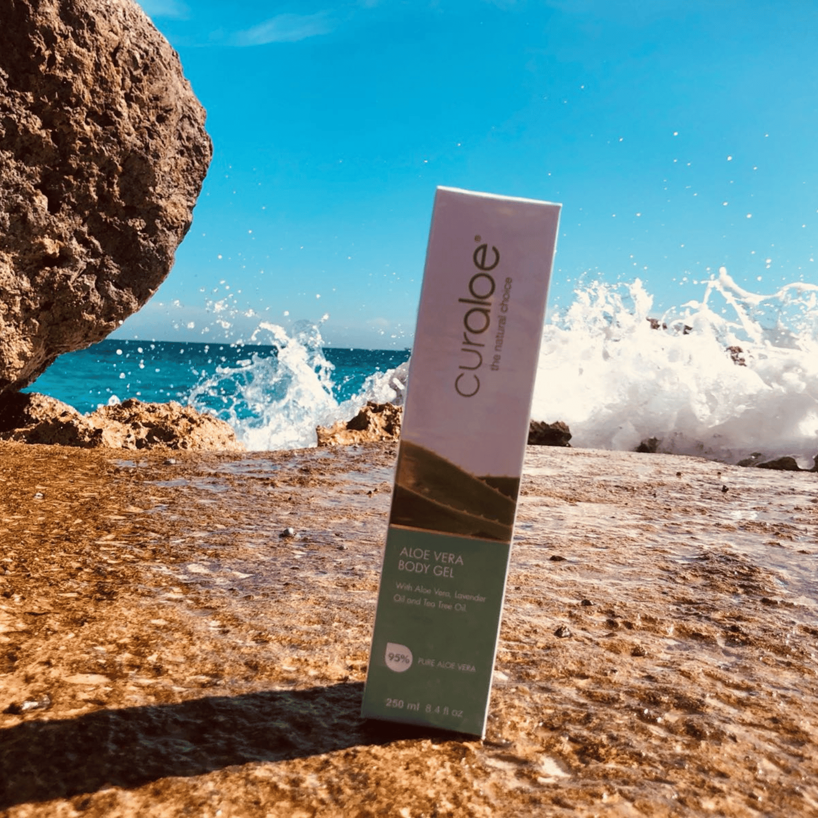 Curaloe Aloe Vera Body Gel packaging on a rocky beach with ocean waves.