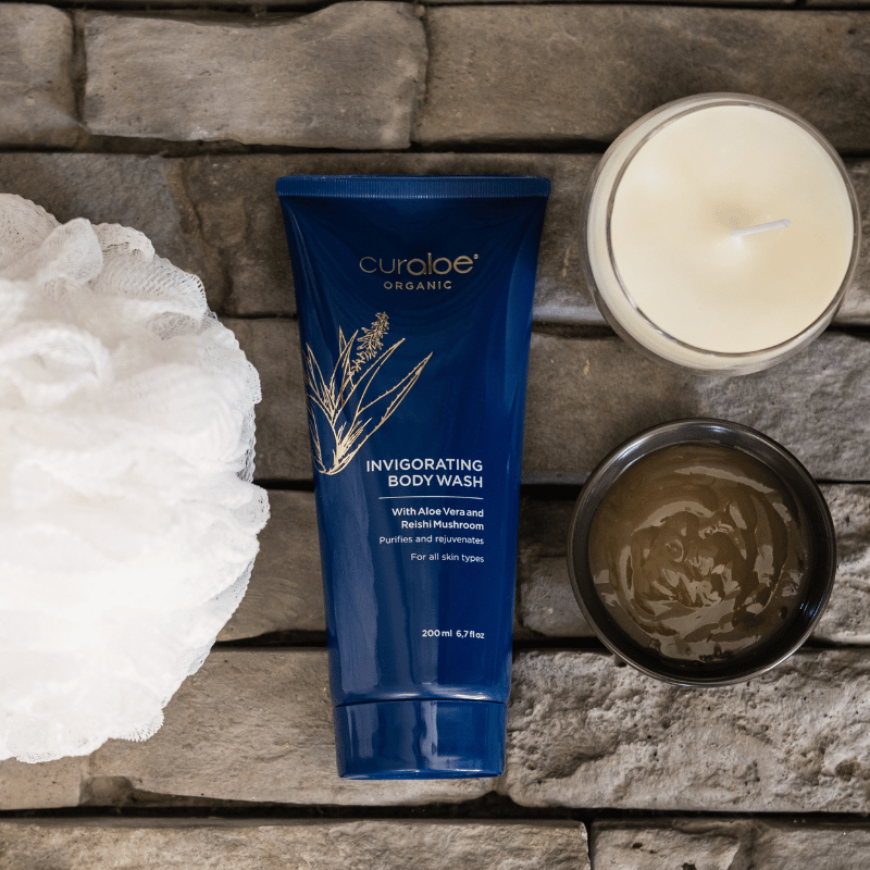 Curaloe Organic Invigorating Body Wash tube on a stone surface with a candle and loofah.