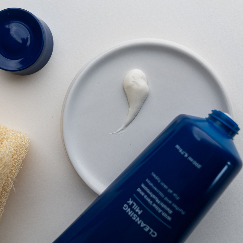 Blue bottle of cleansing milk with a droplet on a white plate, sponge, and blue container in the background.