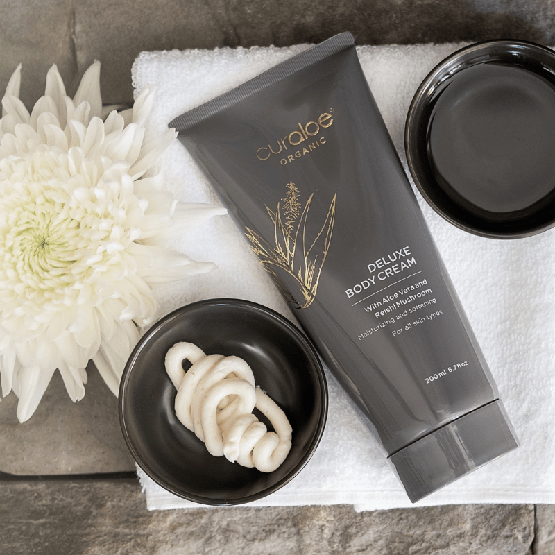Curaloe Organic Deluxe Body Cream with a white flower on a stone surface
