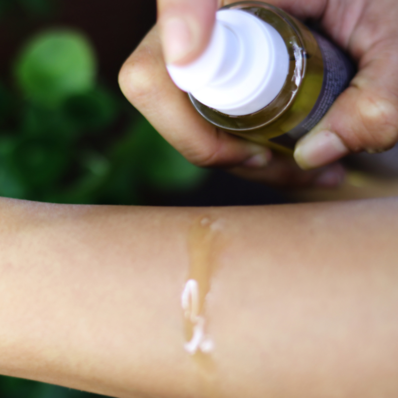 Hand applying a Curaloe Face & Body Oil to a person's arm with a green blurred background