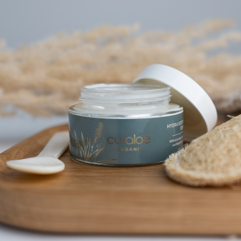 Jar of Curaloe Hydra Restore Cream with a white lid on a wooden board, surrounded by natural elements.