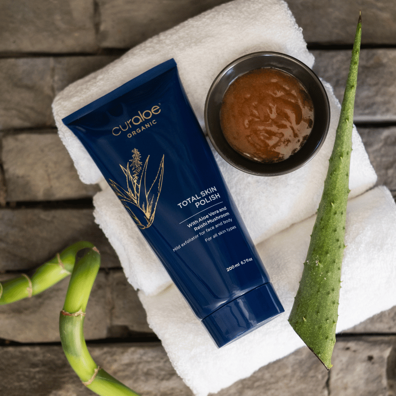 Curaloe Organic Total Skin Polish tube on a towel with aloe vera leaves on a wooden surface