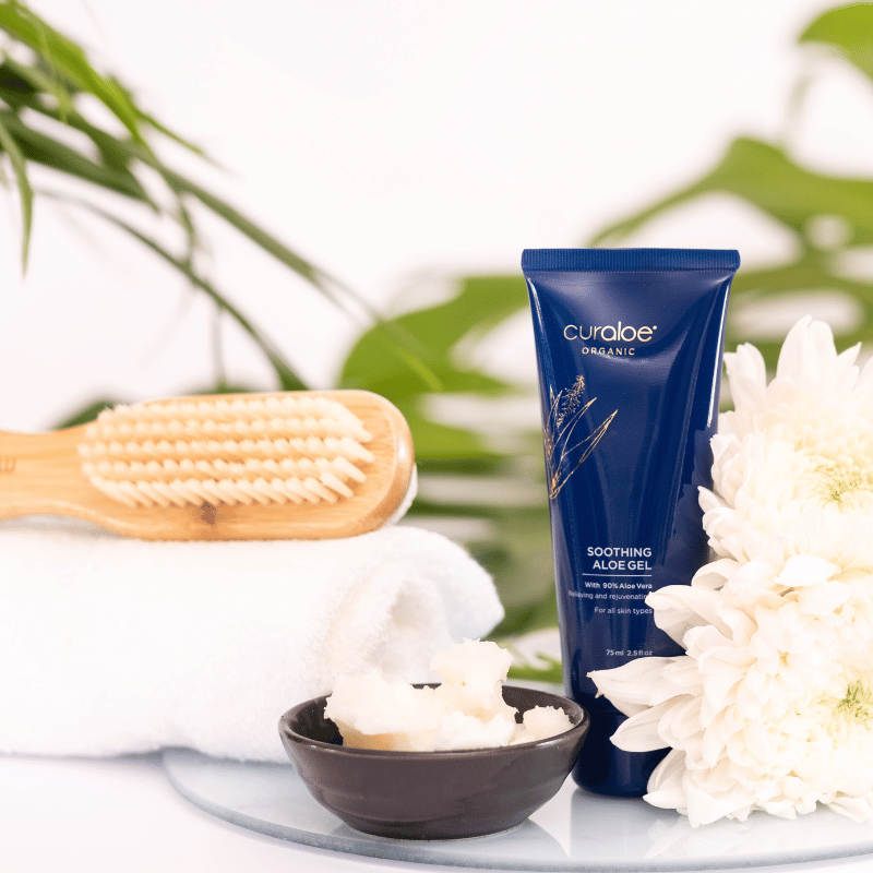Curaloe aloe gel tube with a wooden brush, towel, and flowers on a white surface.