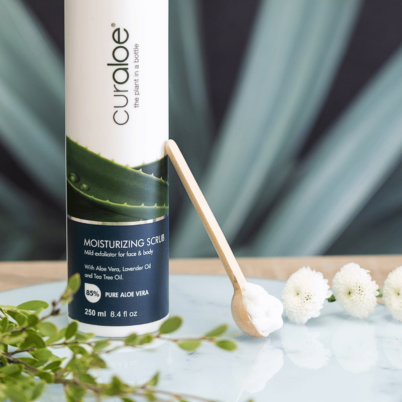 Curaloe moisturizing scrub bottle with a wooden spoon and flowers on a light blue surface.