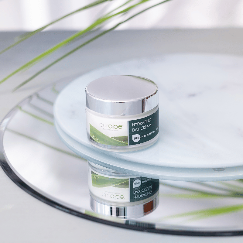 Cylindrical jar of Curoloe Hydrating Day Cream on a reflective surface with green leaves in the background